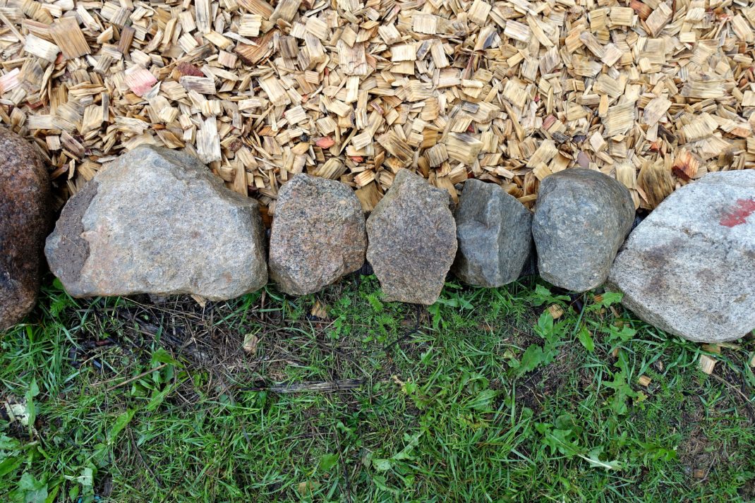 En kant av stora grå stenar i mitten av bilden, träflis i övre delen och gräs nedanför kanten. Edge your beds, a row of rocks edging my beds