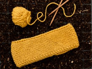 Ett gult stickat pannband bredvis ett par stickor och ett garnnystan